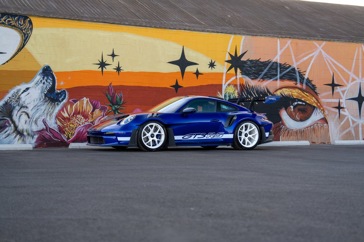Blue 2025 Porsche 911 GT3 RS with white livery and white rims parked in front of a mural.