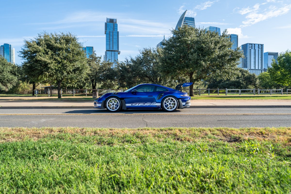 A blue 2025 Porsche 911 GT3 RS with white center-lock rims and "GT3 RS" livery drives past city buildings.