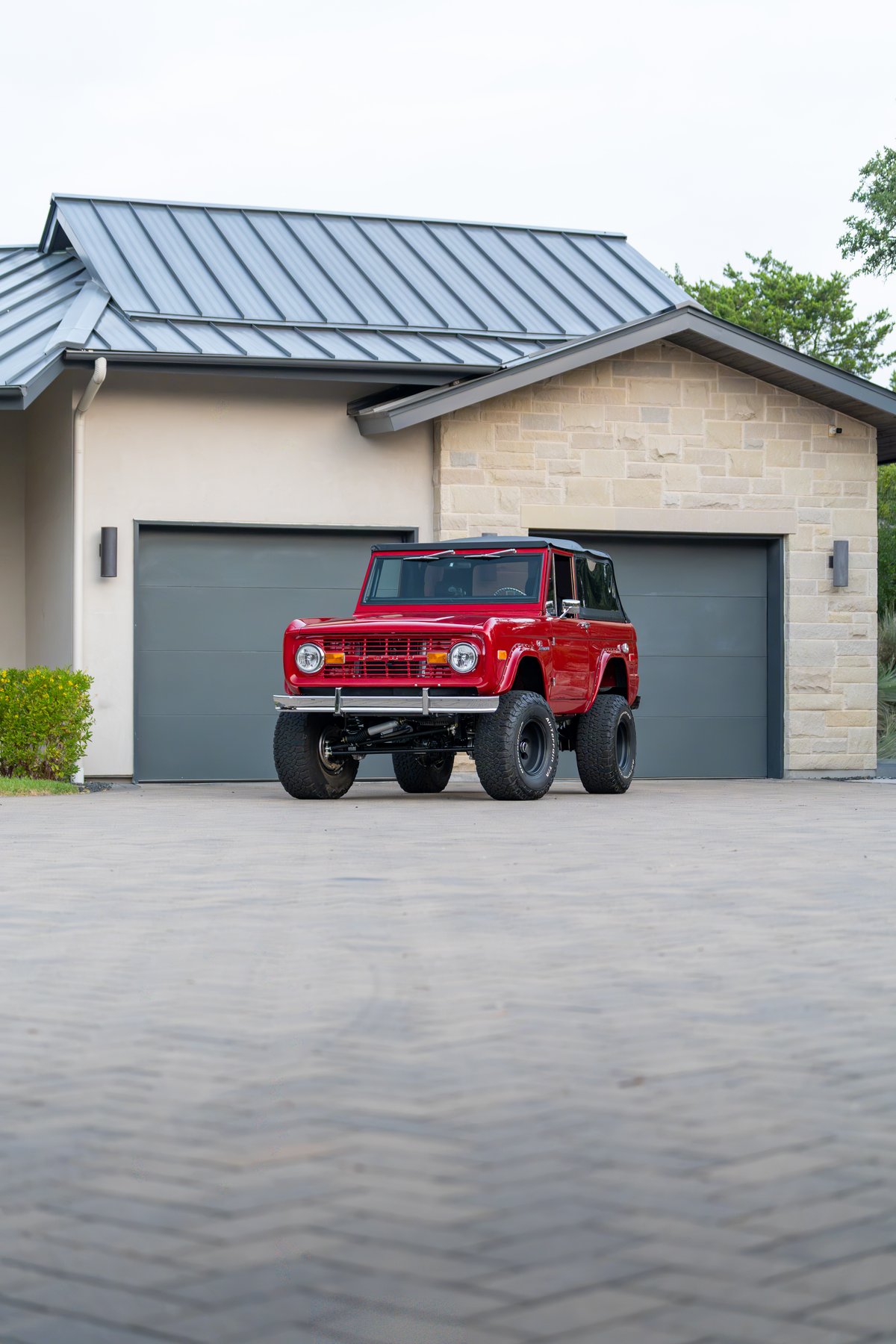 Red 1966 Ford Bronco with black rims and lift kit parked in front of a modern home.