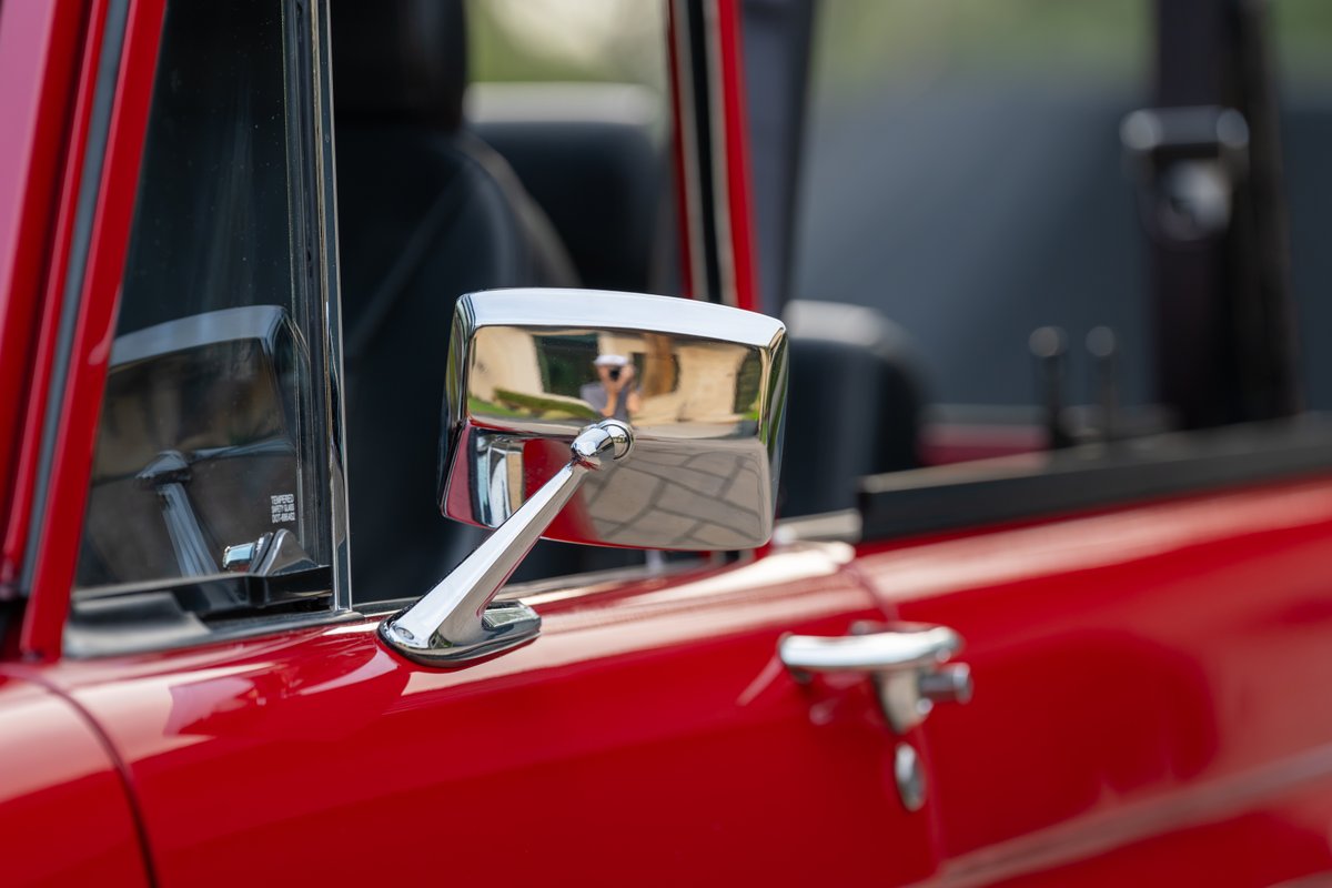 Close-up of a red 1966 Ford Bronco's chrome side mirror and door handle.