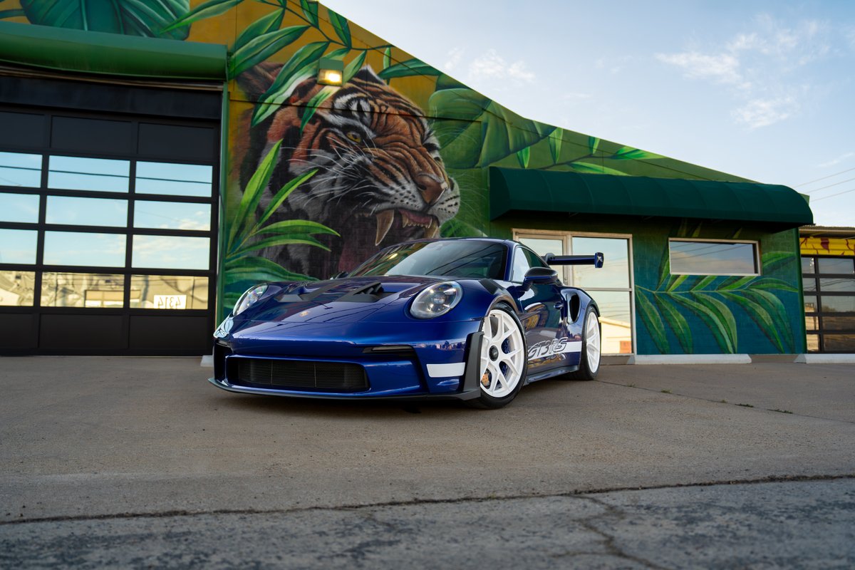 Blue 2025 Porsche 911 GT3 RS with white racing livery & rims, parked outside a mural.