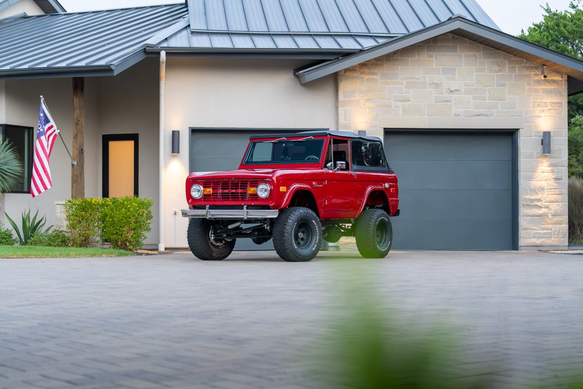 Red 1966 Ford Bronco with black rims and off-road tires parked in front of a modern home.
