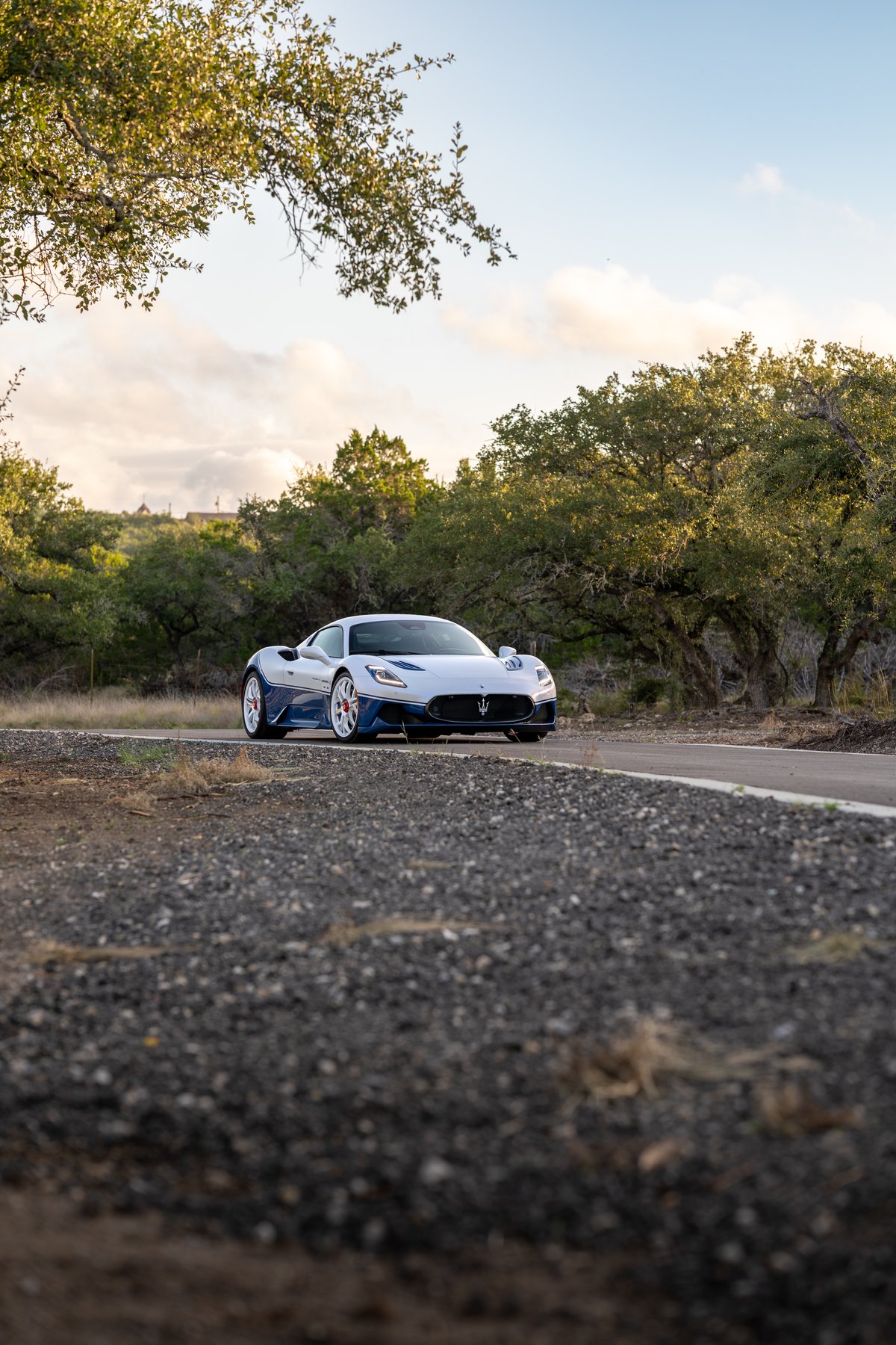 2024 Maserati MC20 Icona in blue and white with white rims driving on a rural road.