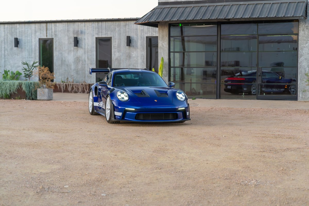 A blue 2025 Porsche 911 GT3 RS with white rims and a prominent rear wing sits parked on a dirt lot.