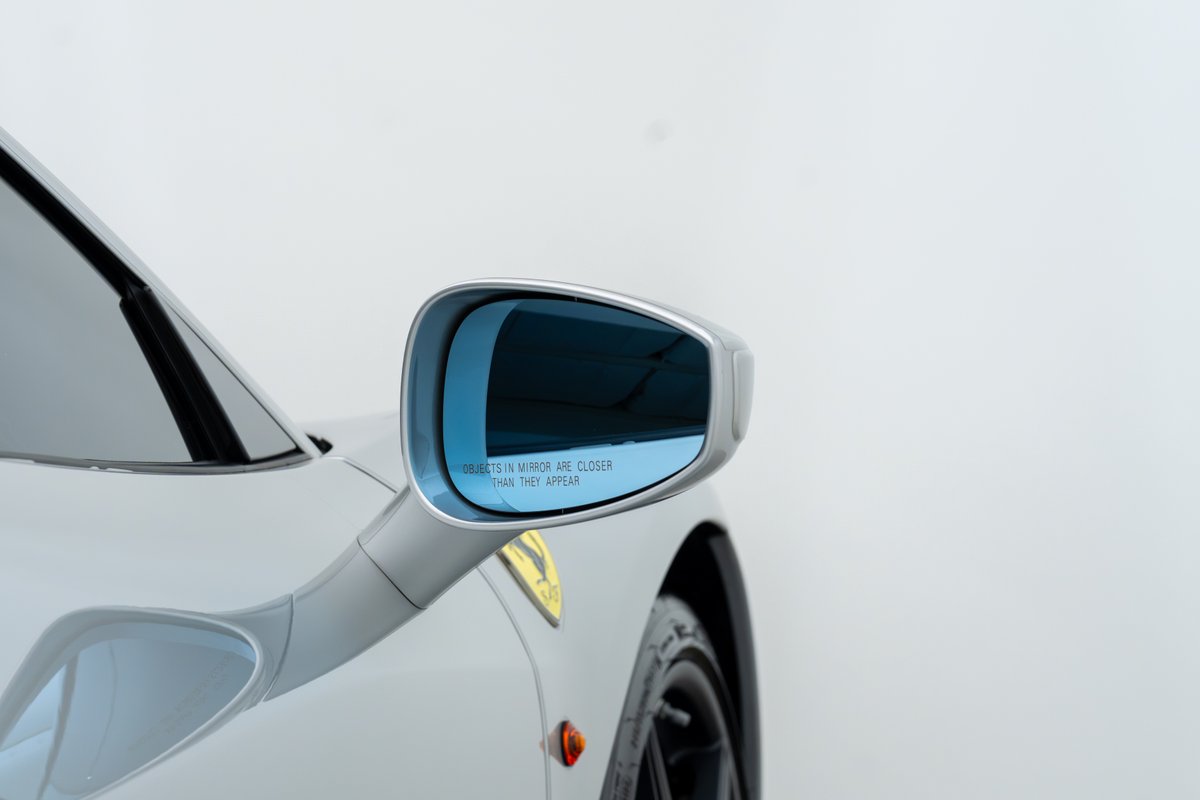 Close-up of a silver 2015 Ferrari 458 Speciale side mirror with blue tint and yellow shield emblem.