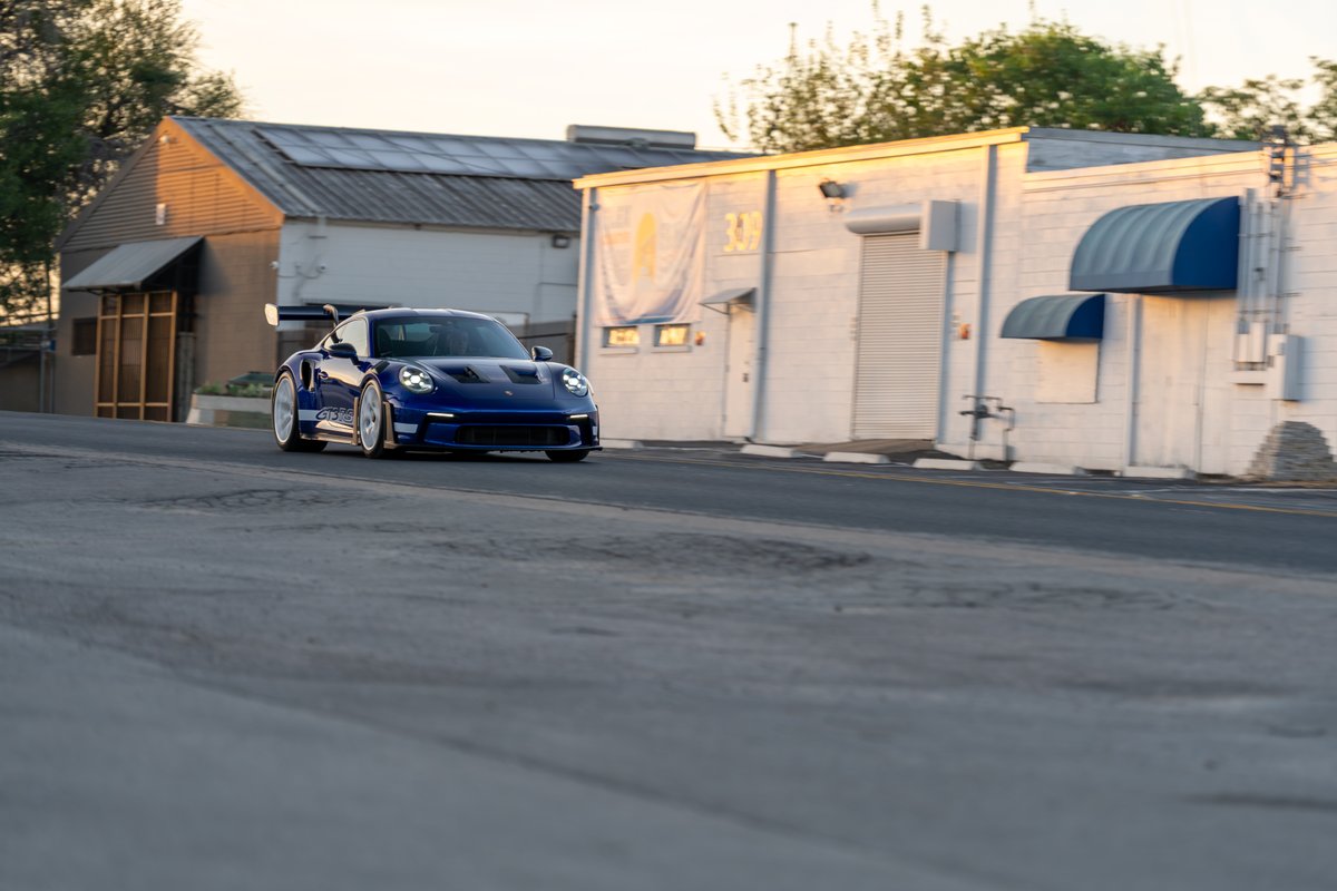 A 2025 blue Porsche 911 GT3 RS with white rims and GT3 RS livery drives on a road.