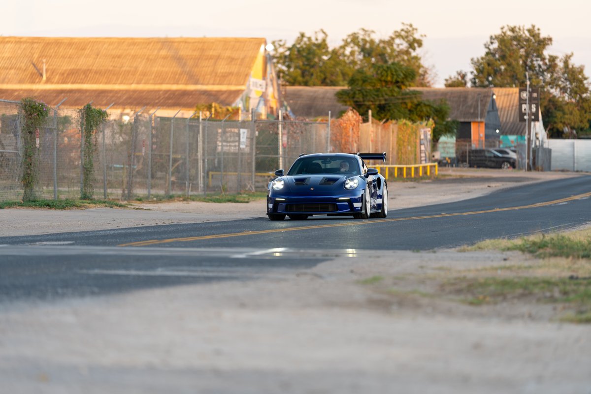 A blue 2025 Porsche 911 GT3 RS with a large rear wing and silver rims drives on a road.