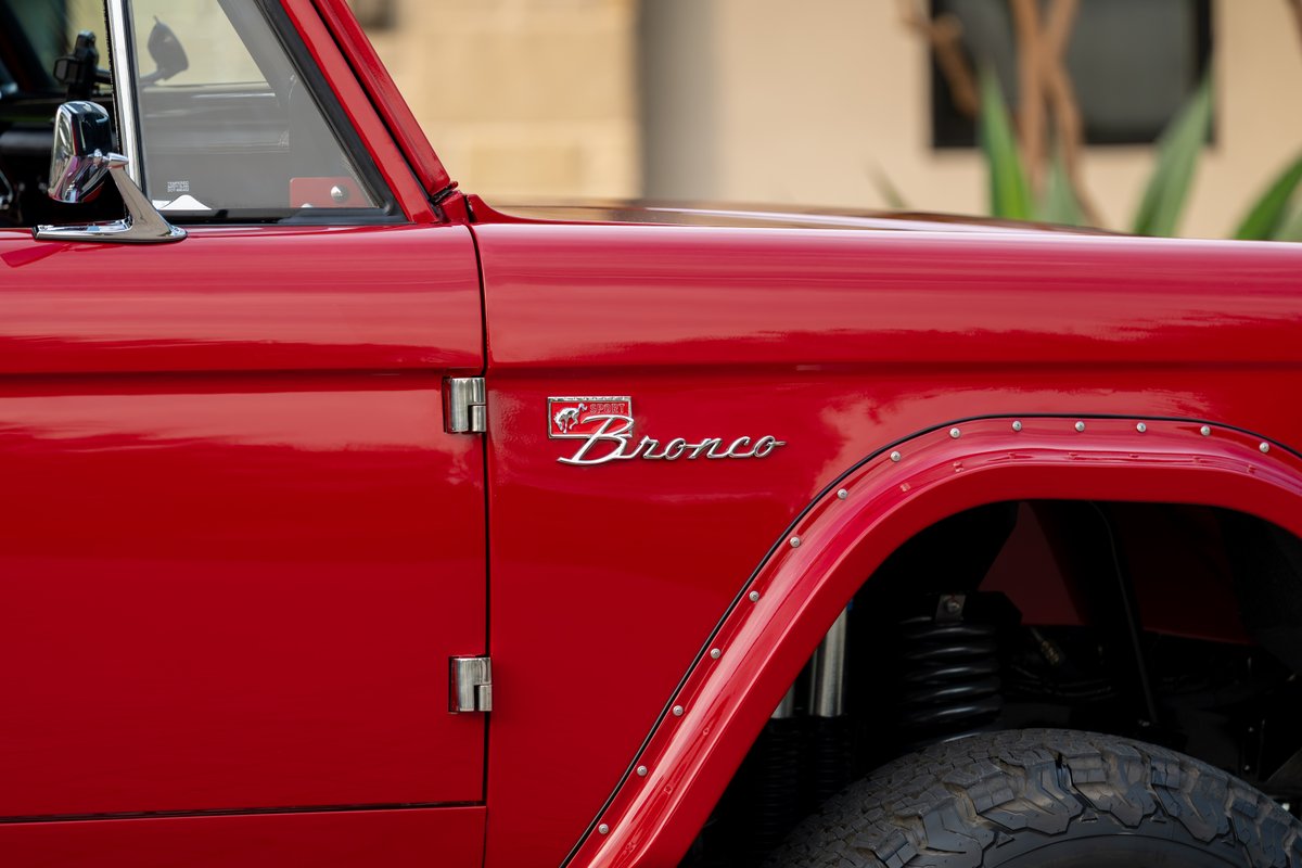 Close-up of a red 1966 Ford Bronco with a chrome "Bronco" badge and riveted fender flares.
