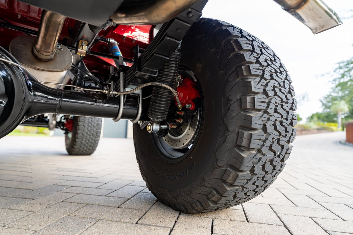 Close-up of a red 1966 Ford Bronco's rear axle, leaf springs, and off-road tire. Red brake calipers visible.