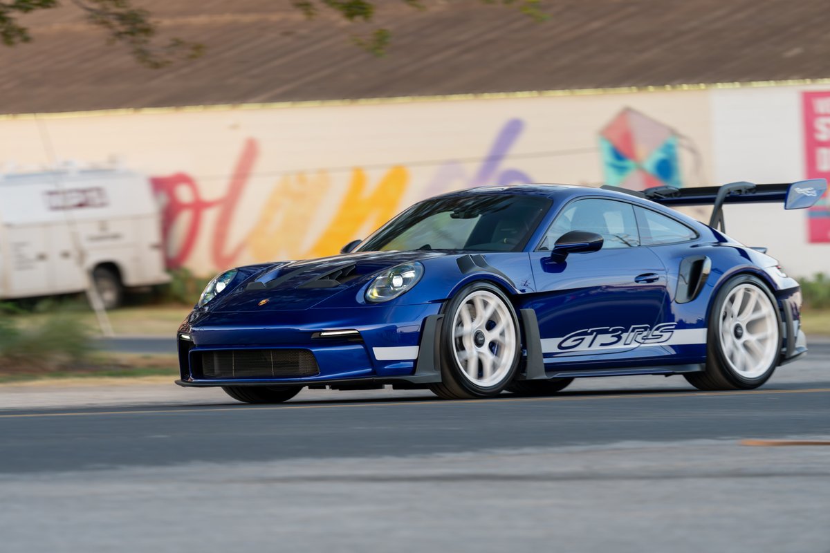 A blue 2025 Porsche 911 GT3 RS with white GT3 RS livery and white center-lock rims drives on a road.