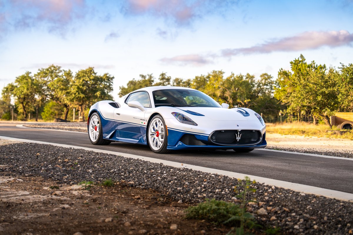 A 2024 Maserati MC20 Icona, white with blue livery and white rims, drives on a paved road.