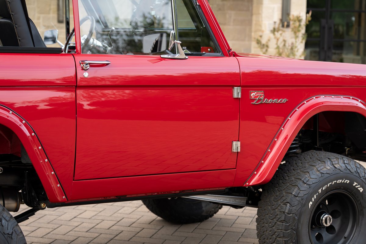Red 1966 Ford Bronco by Atlas Broncos. Chrome door handle, fender flares, and "Bronco" emblem. Black rims with All-Terrain tires.