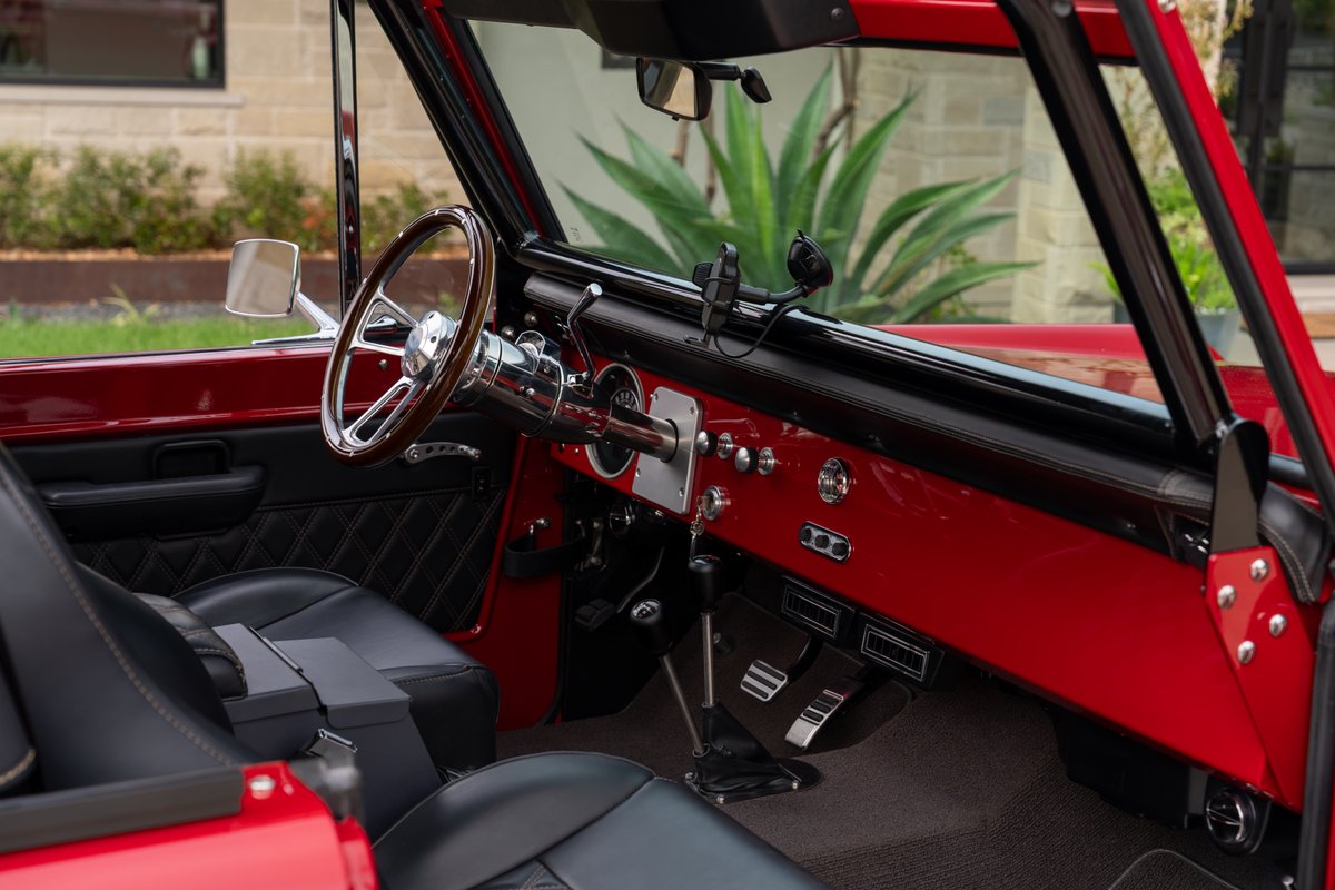 1966 Ford Bronco interior with red dash, wood steering wheel, and black diamond-stitched seats.