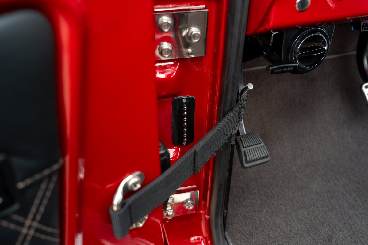 Red 1966 Ford Bronco door jamb with a black fabric strap, speaker grille, and floor pedals.