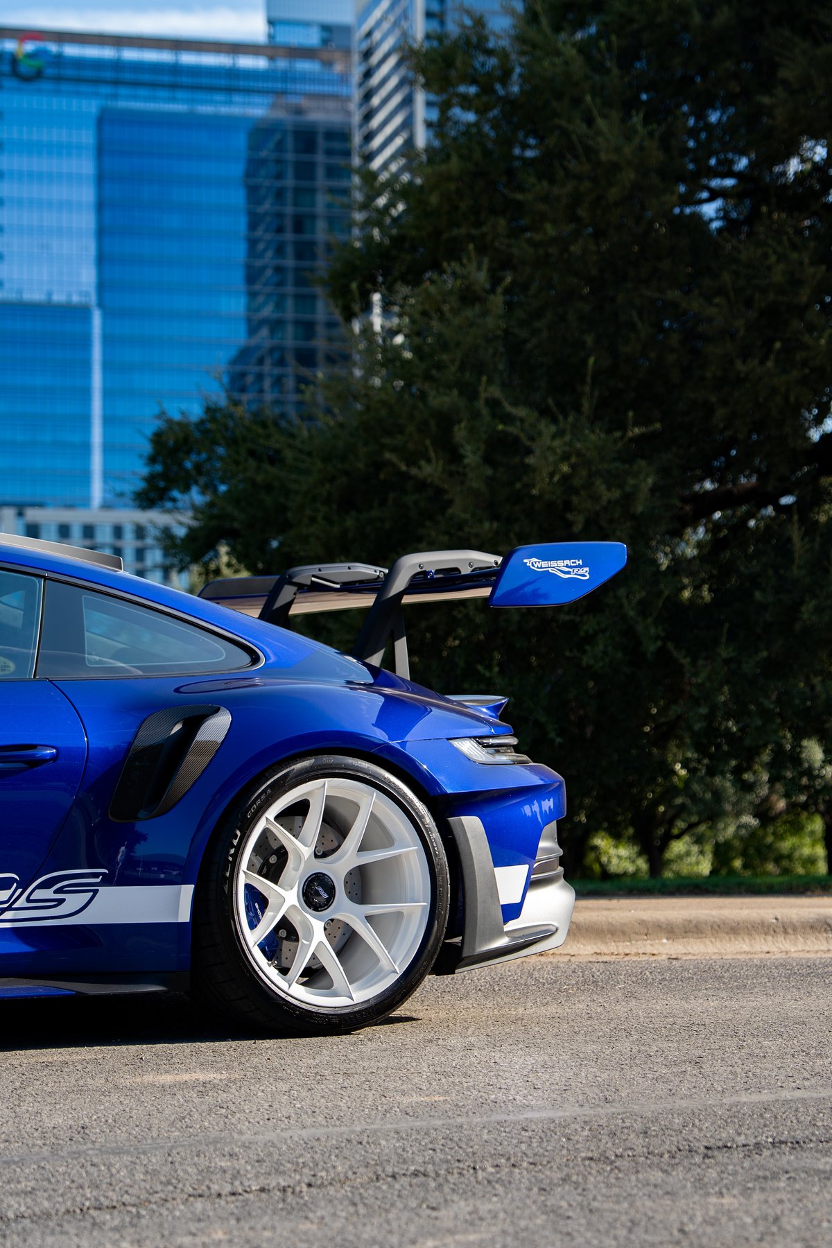 2025 Porsche 911 GT3 RS in blue with white racing livery, white rims, and Weissach package spoiler.