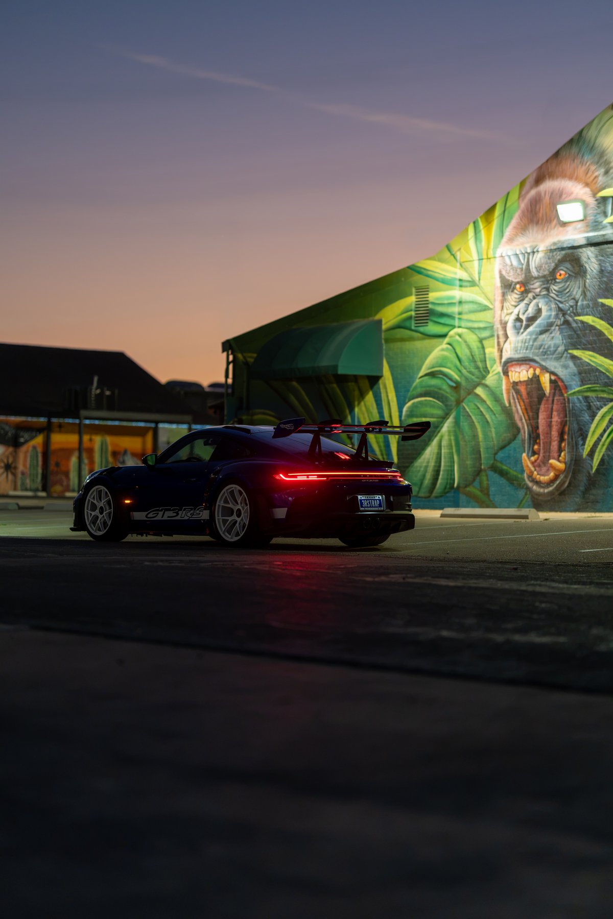 A dark blue 2025 Porsche 911 GT3 RS with white rims and a large rear wing sits parked at dusk.