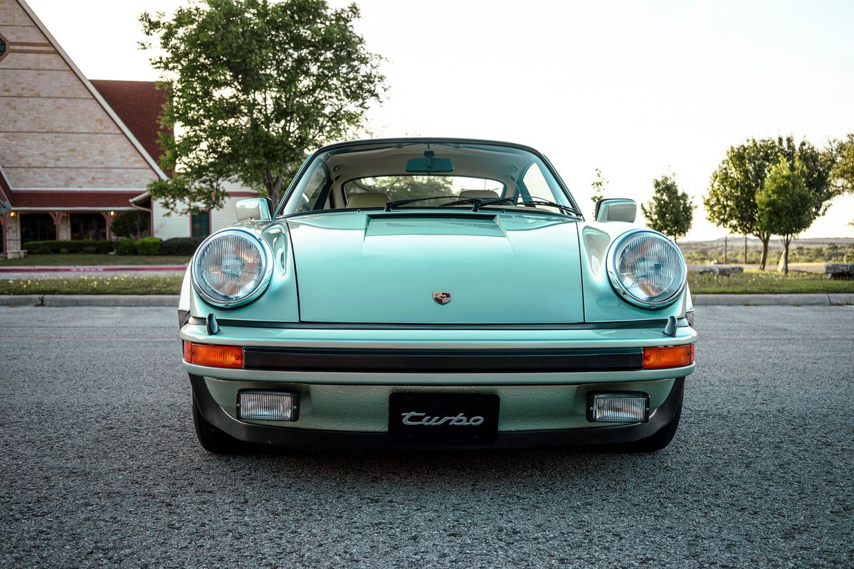 Head-on 1976 Porsche 930 Turbo in Ice Metallic Green. Features widebody fenders, round headlights, and "Turbo" script.