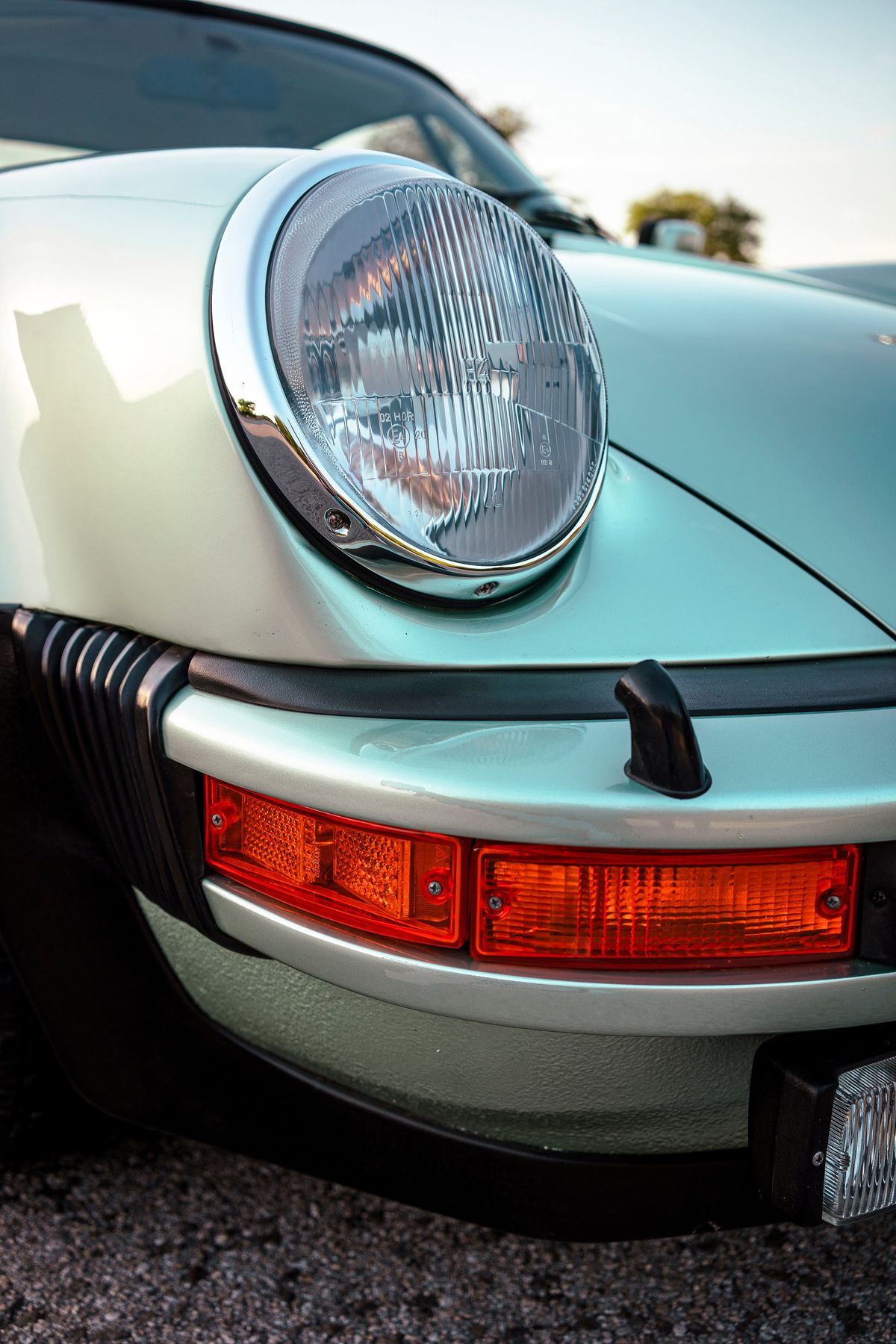 Front detail of an ice metallic green 1976 Porsche 930, highlighting the round headlight with chrome bezel and amber bumper-mounted turn signals.