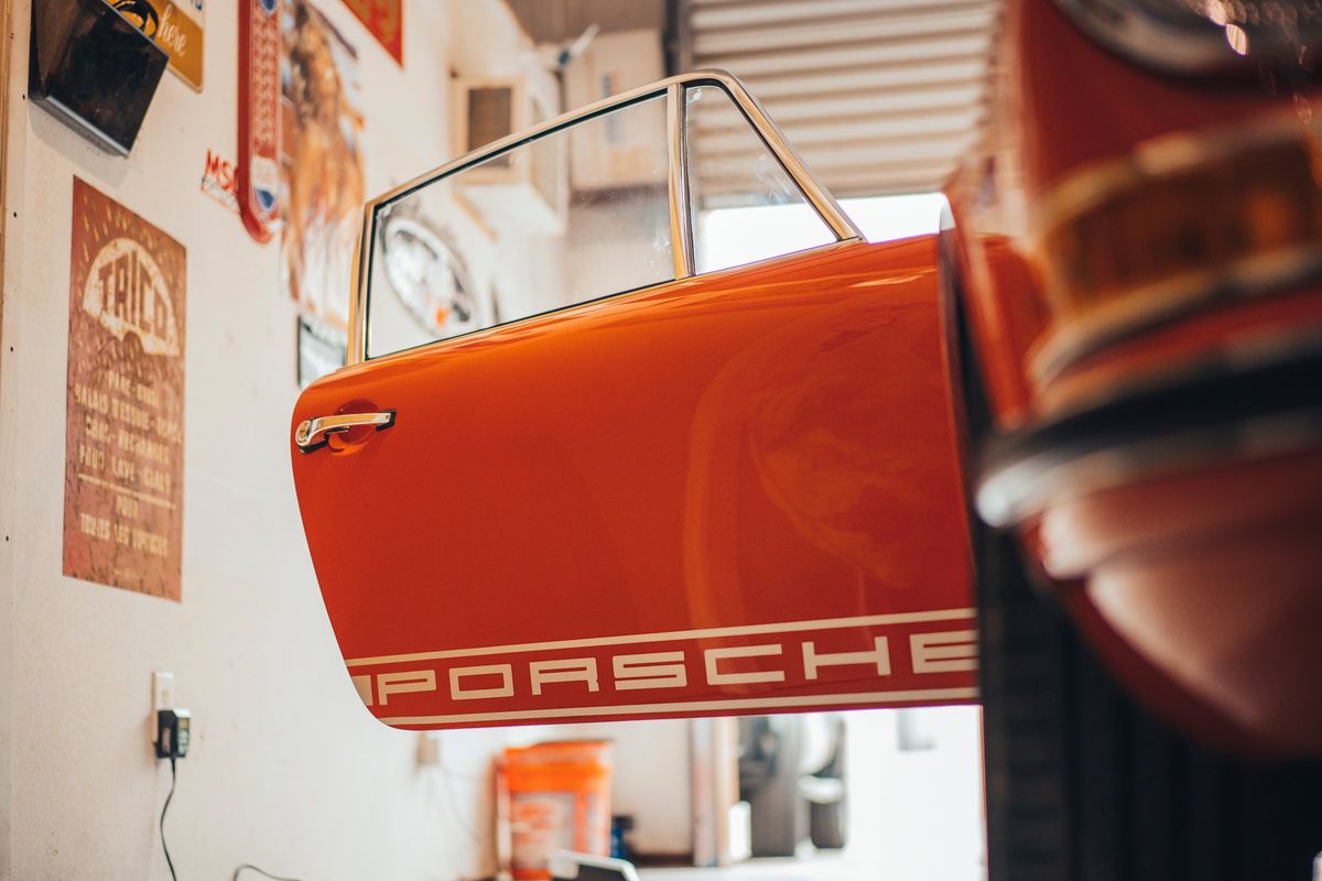 Vintage Tangerine Porsche 911 door, open, featuring white PORSCHE script livery, chrome window frame & handle in a garage.