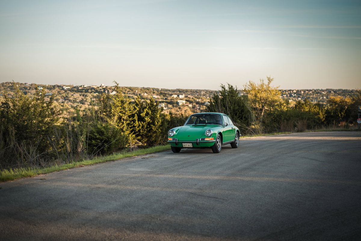 Pristine Conda Green 1971 Porsche 911E coupe with chrome trim and Fuchs-style alloy rims on a scenic road.