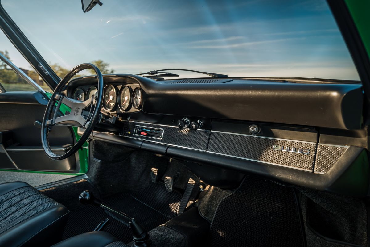 1971 Porsche 911E interior. Driver's view of black dashboard, iconic five-gauge cluster, four-spoke wheel, manual shifter, and textured seats.