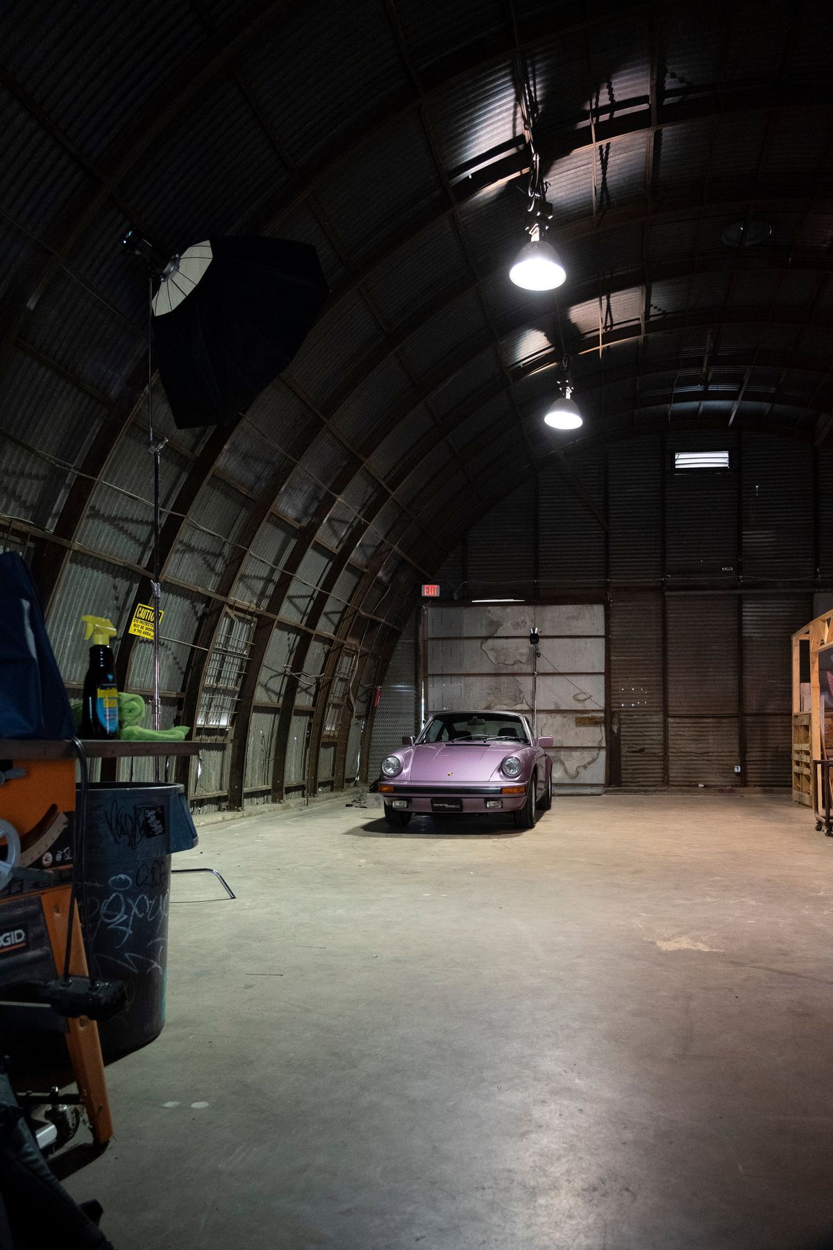 Front view: Metallic purple Porsche 911 G-body coupe with black impact bumpers & Fuchs-style rims in a warehouse.
