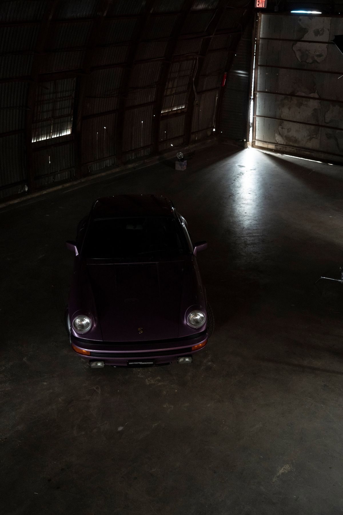 Purple Porsche 911 G-series coupe. Overhead shot showcasing classic round headlights & black bumper trim in dark industrial space.