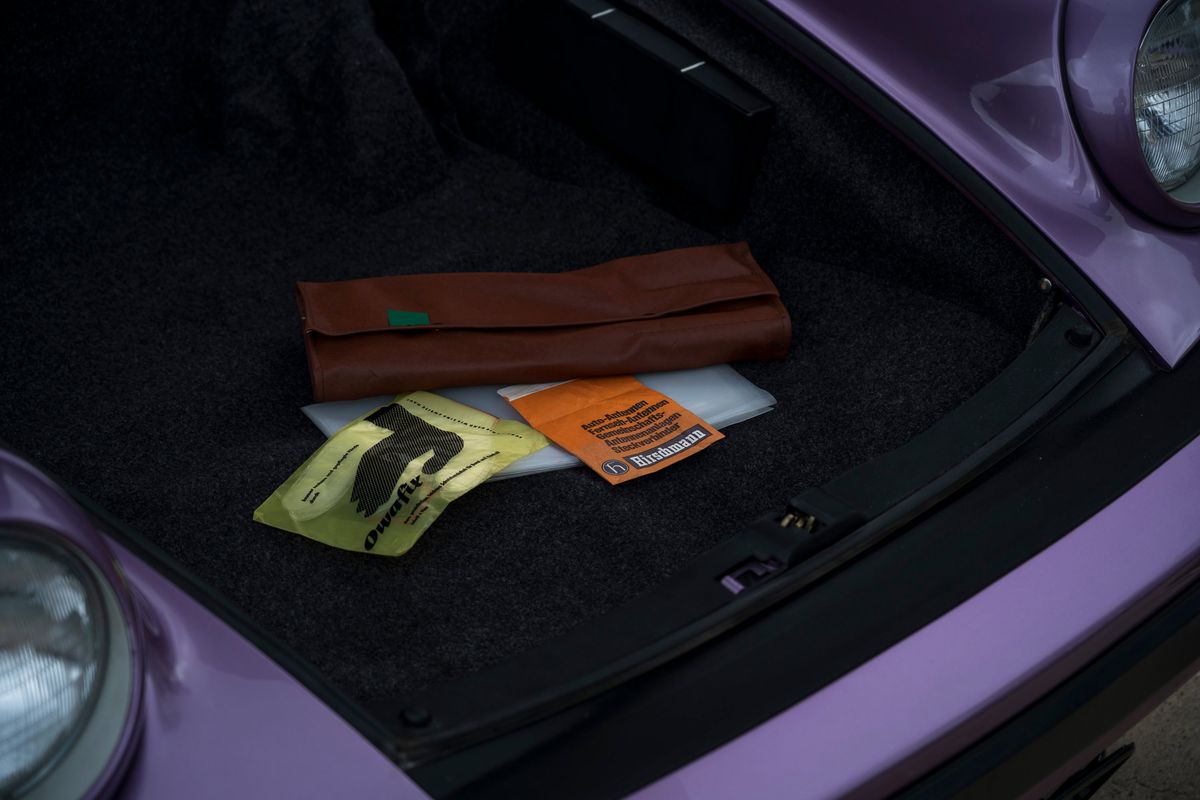 Close-up of a purple Porsche's frunk: carpeted interior with a vintage brown tool roll, Owafix, and Hirschmann antenna component.