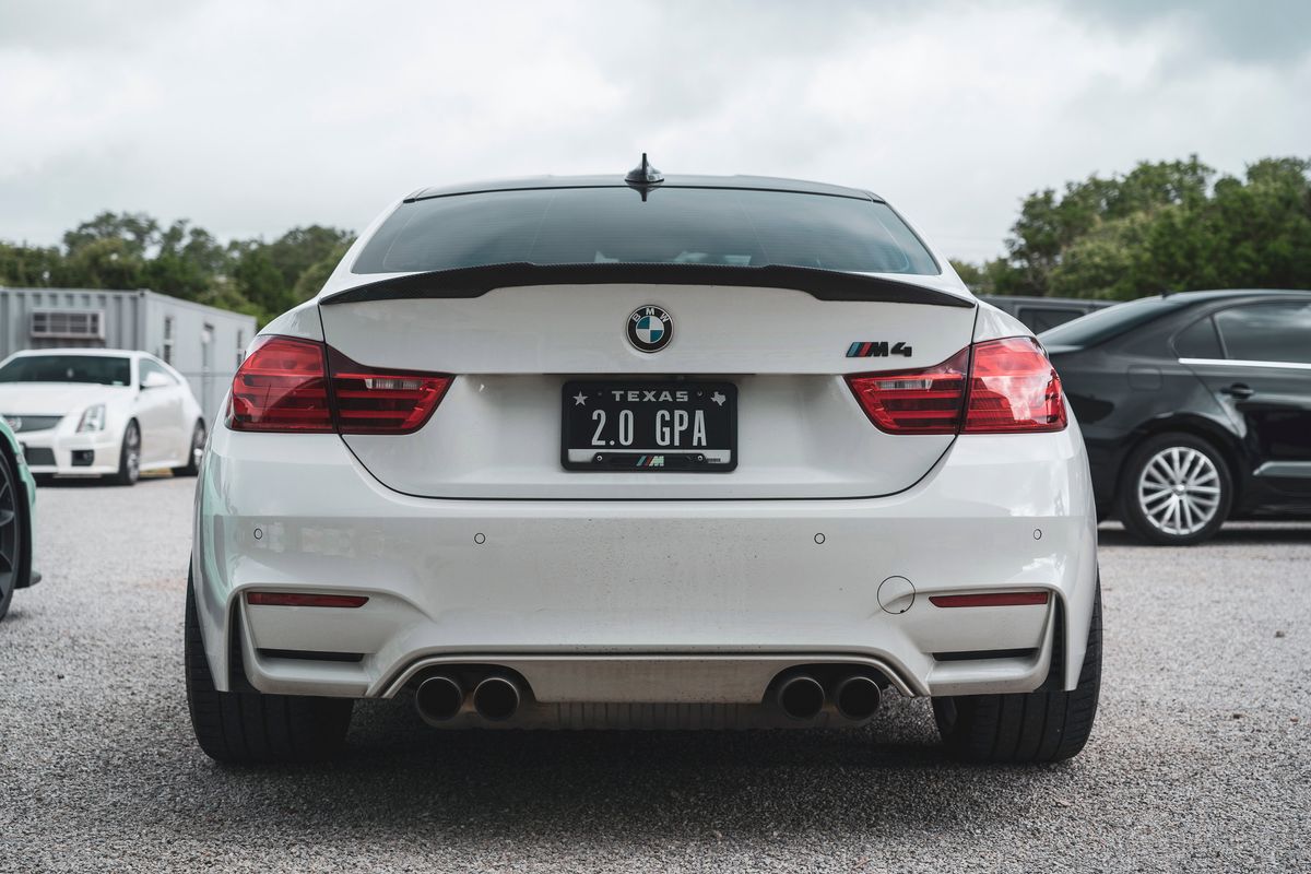 White BMW M4 (F82) rear with carbon fiber trunk spoiler, quad exhausts, and a Texas "2.0 GPA" license plate.