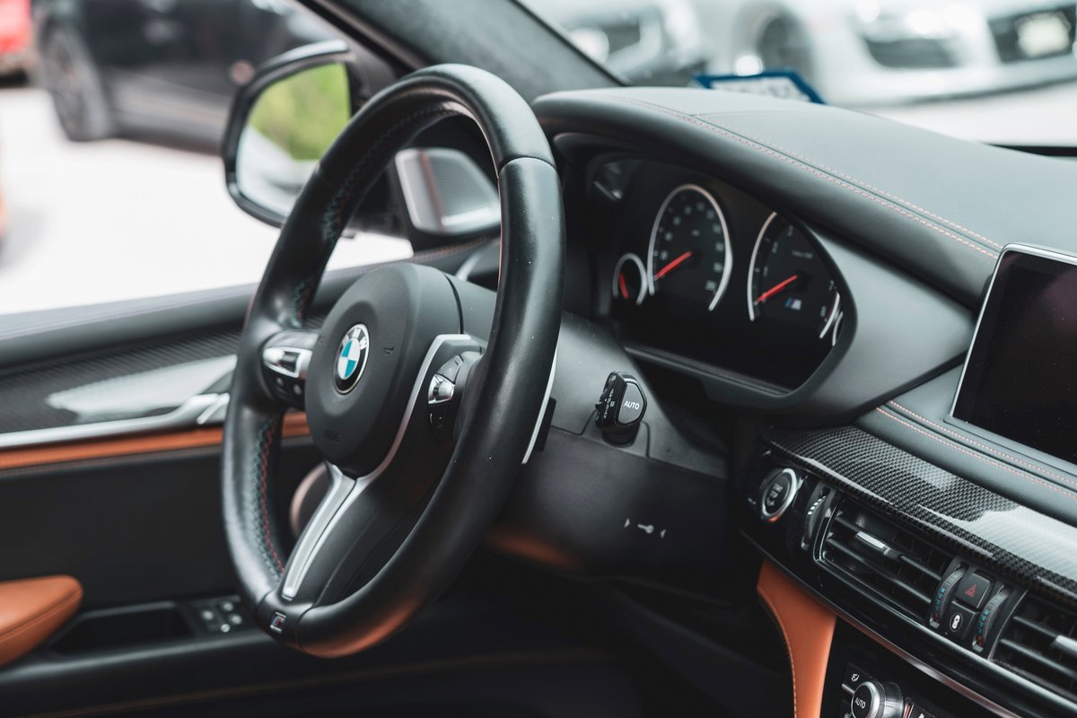 BMW X6 M (F86) interior: M leather steering wheel, carbon fiber dashboard trim, saddle brown leather accents, and red contrast stitching.