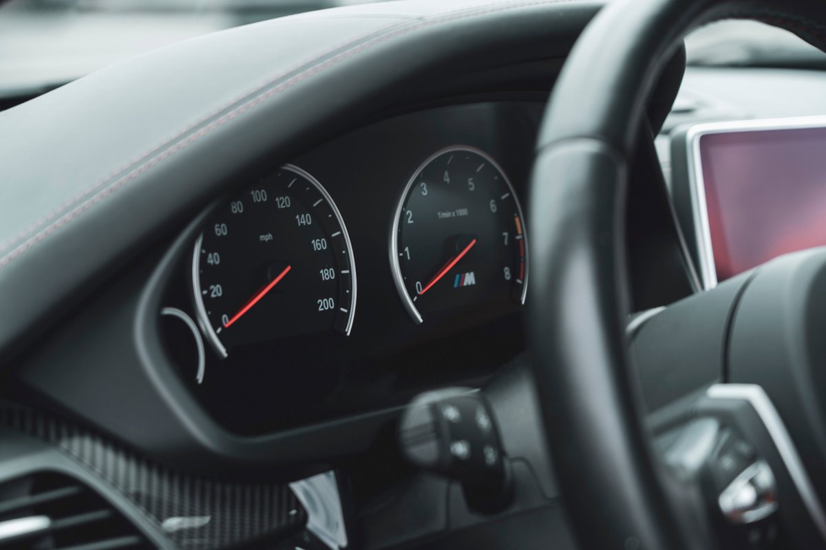 BMW M dashboard view: carbon fiber trim, red stitching, M-badged tachometer and speedometer. Modern sports car interior.