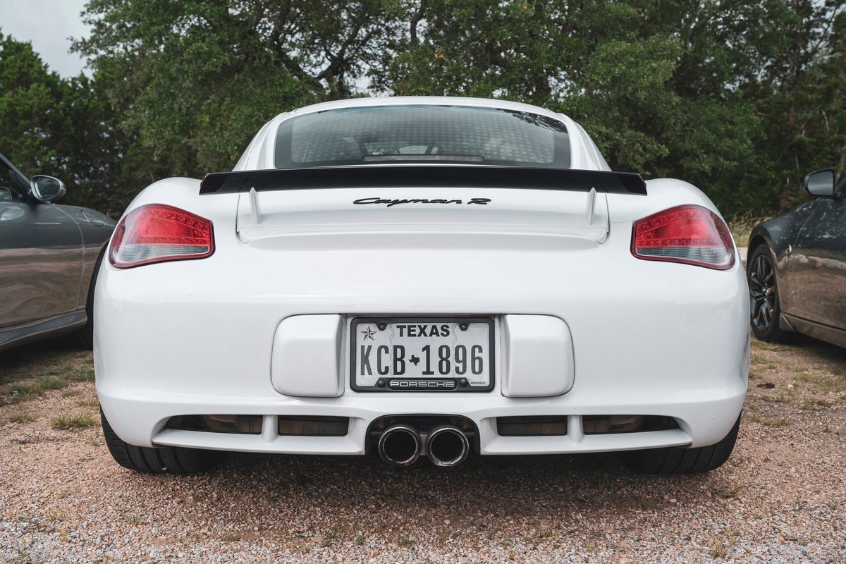White Porsche Cayman R rear view, featuring black fixed spoiler, dual chrome exhaust, and bright LED taillights at a car meet.
