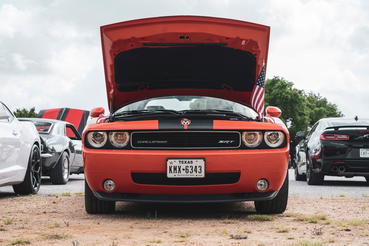 Front view of an orange Dodge Challenger SRT, hood open, showcasing black racing stripes, grille with SRT badge, and fog lights.