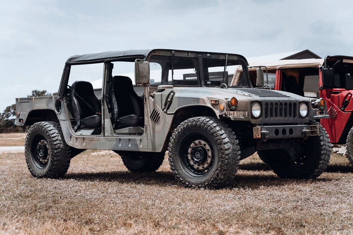 Military-style Hummer H1 with olive drab camo livery, soft top, and no doors. Features mud-terrain tires on black beadlock rims.