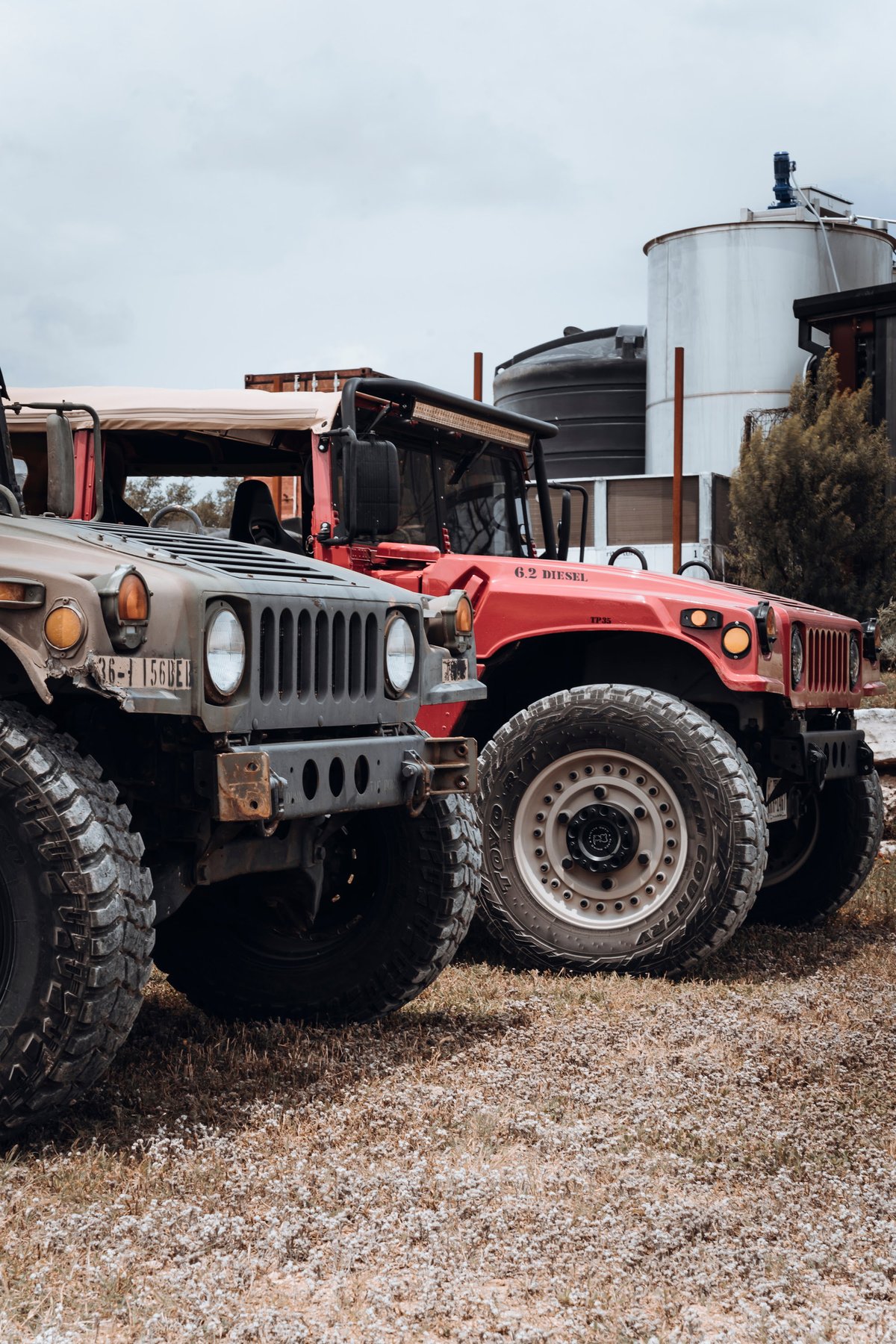 Green military HMMWV with off-road tires & a red Hummer H1 6.2 Diesel with custom rims, both with soft tops.