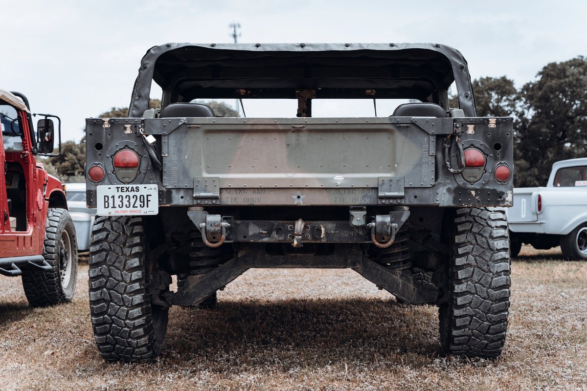 Rear of an olive drab military HMMWV (Humvee) with a soft top & rugged mud-terrain tires. Features heavy-duty recovery points.