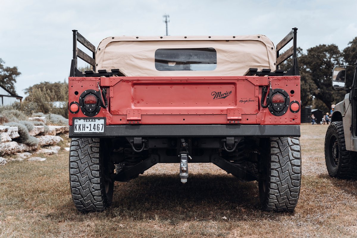 Texas-plated red Hummer H1 rear, tan canvas soft top, "Merica" tailgate, LED taillights, rugged off-road tires.