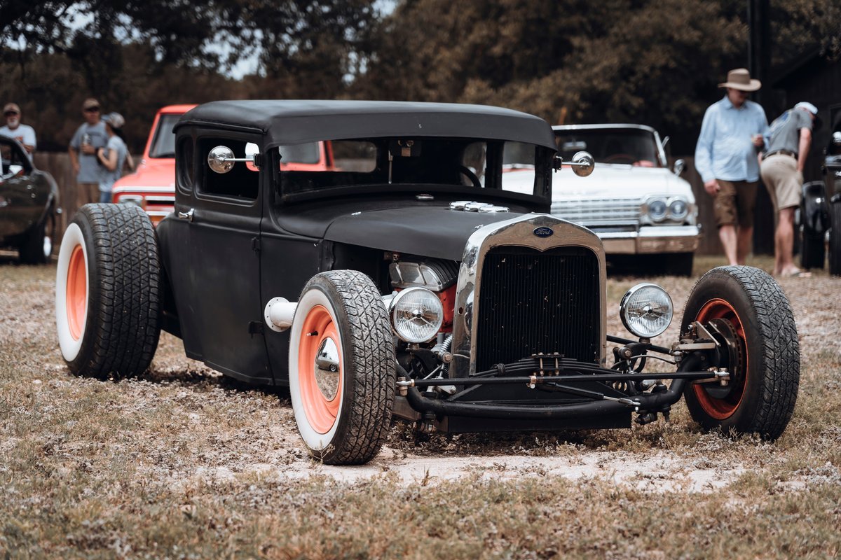 Matte black Ford Model A hot rod with chopped roof, visible V8, front white wall tires, and vibrant orange rims.