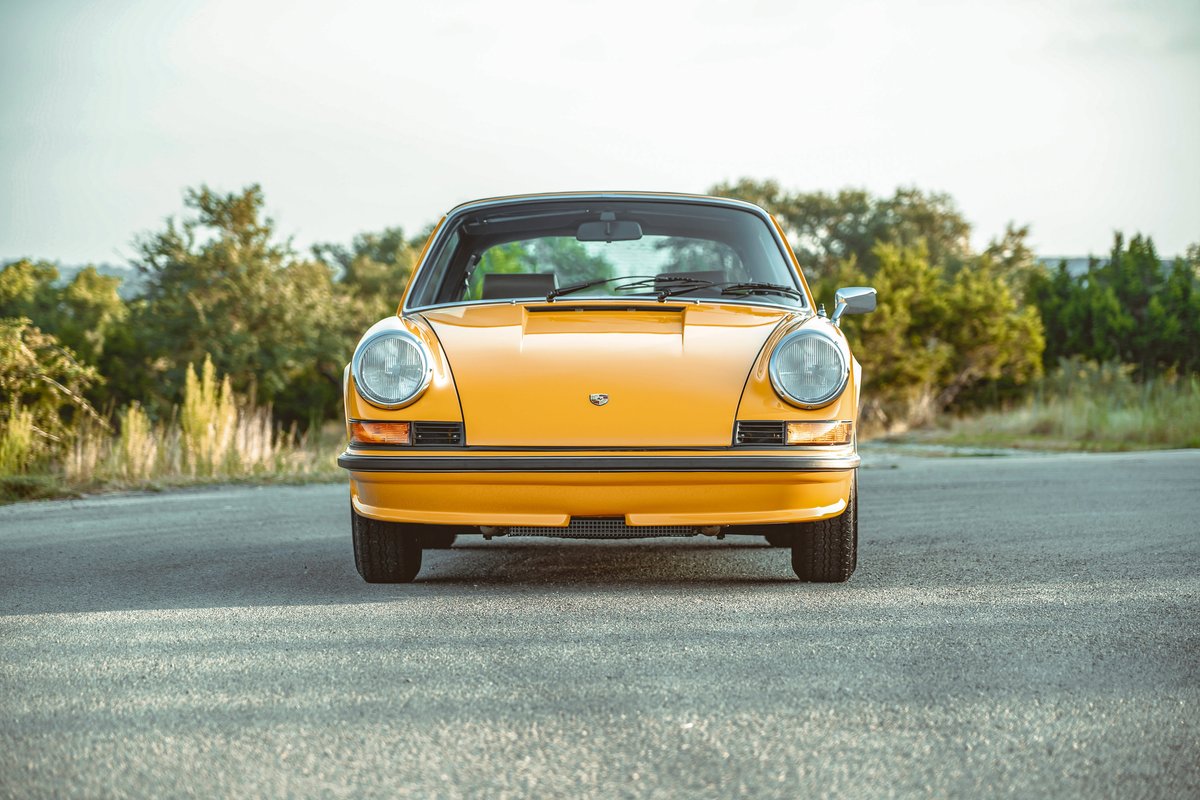 Front view of a 1973 Signal Yellow Porsche 911S, showcasing iconic round headlights, black bumper trim, and Fuchs wheels.