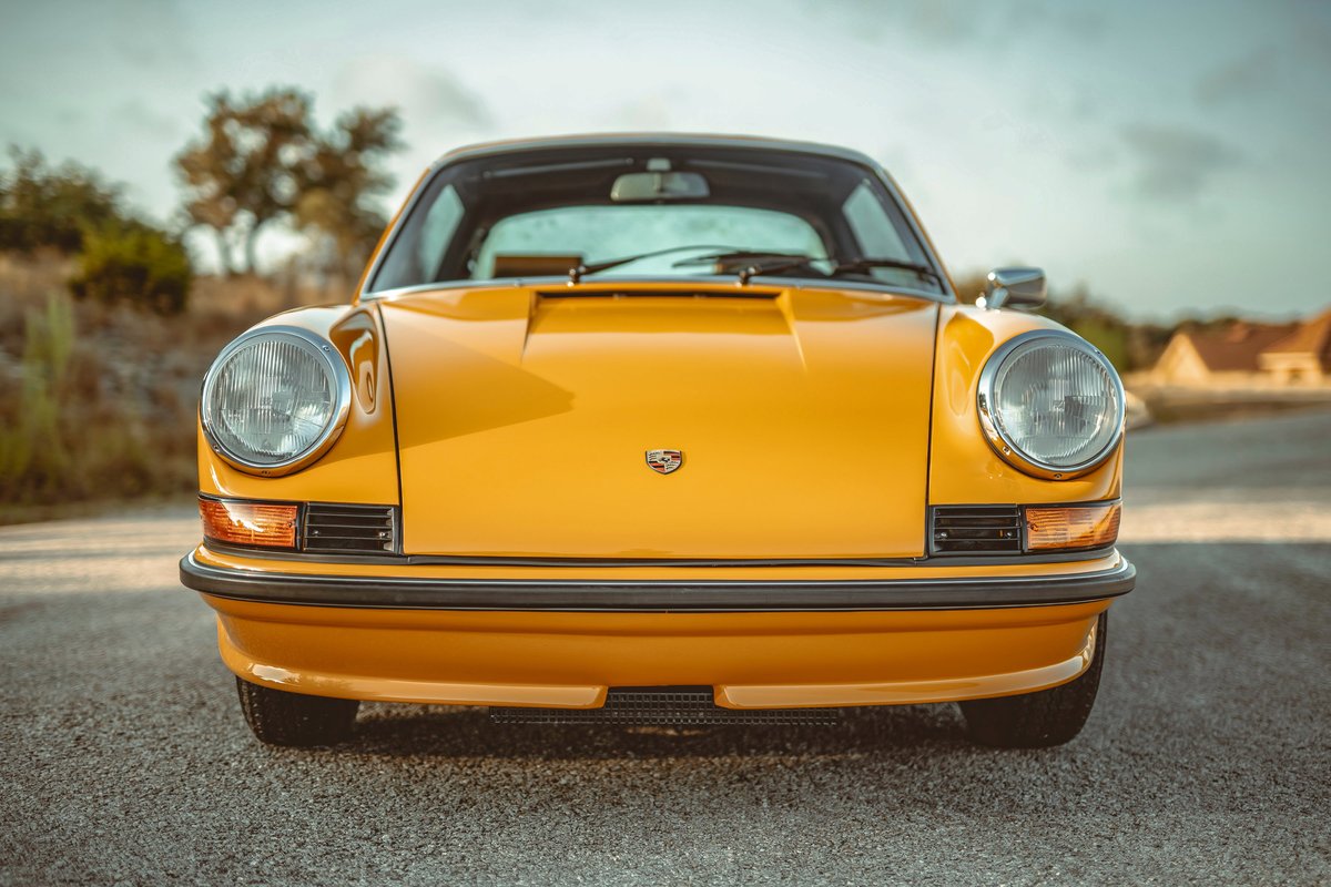 Close-up front view of a Signal Yellow 1973 Porsche 911S, featuring classic round headlights, black bumper trim, and hood crest.