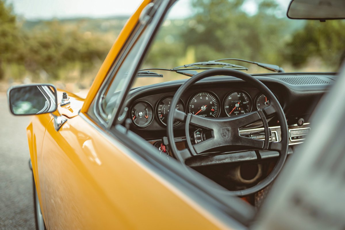Signal Yellow 1973 Porsche 911S, driver's side view. Black interior reveals five-dial instrument cluster and iconic steering wheel.