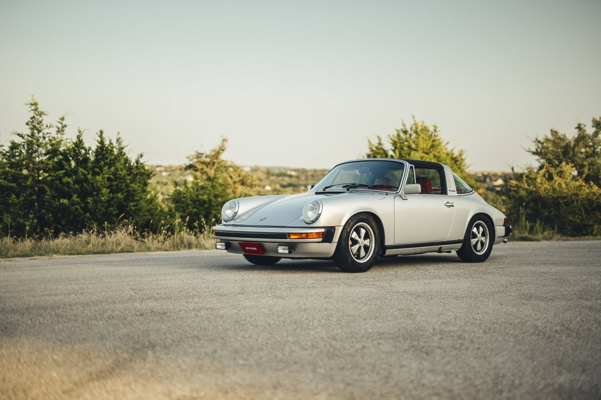 Silver 1977 Porsche 911S Targa with black roof, red interior, classic Fuchs rims, and black trim on an open road.