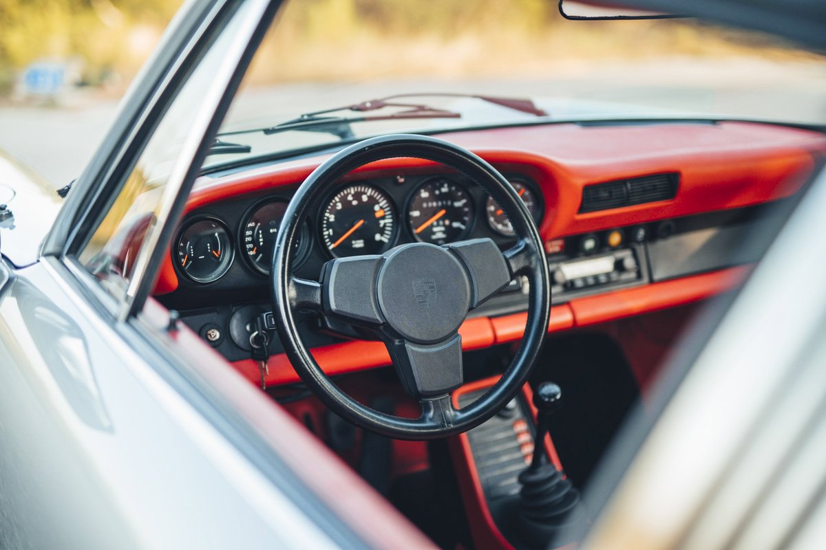 Silver 1977 Porsche 911S interior: red dashboard and trim, black 4-spoke steering wheel, classic gauges, and manual shifter.
