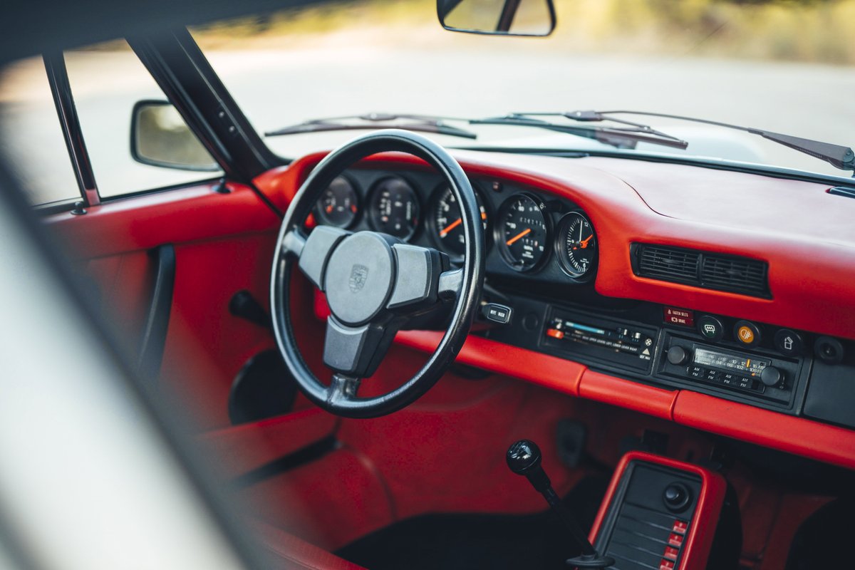 1977 Porsche 911S interior: iconic 5-dial dash, classic steering wheel, and vibrant red trim elements. Manual shifter visible.