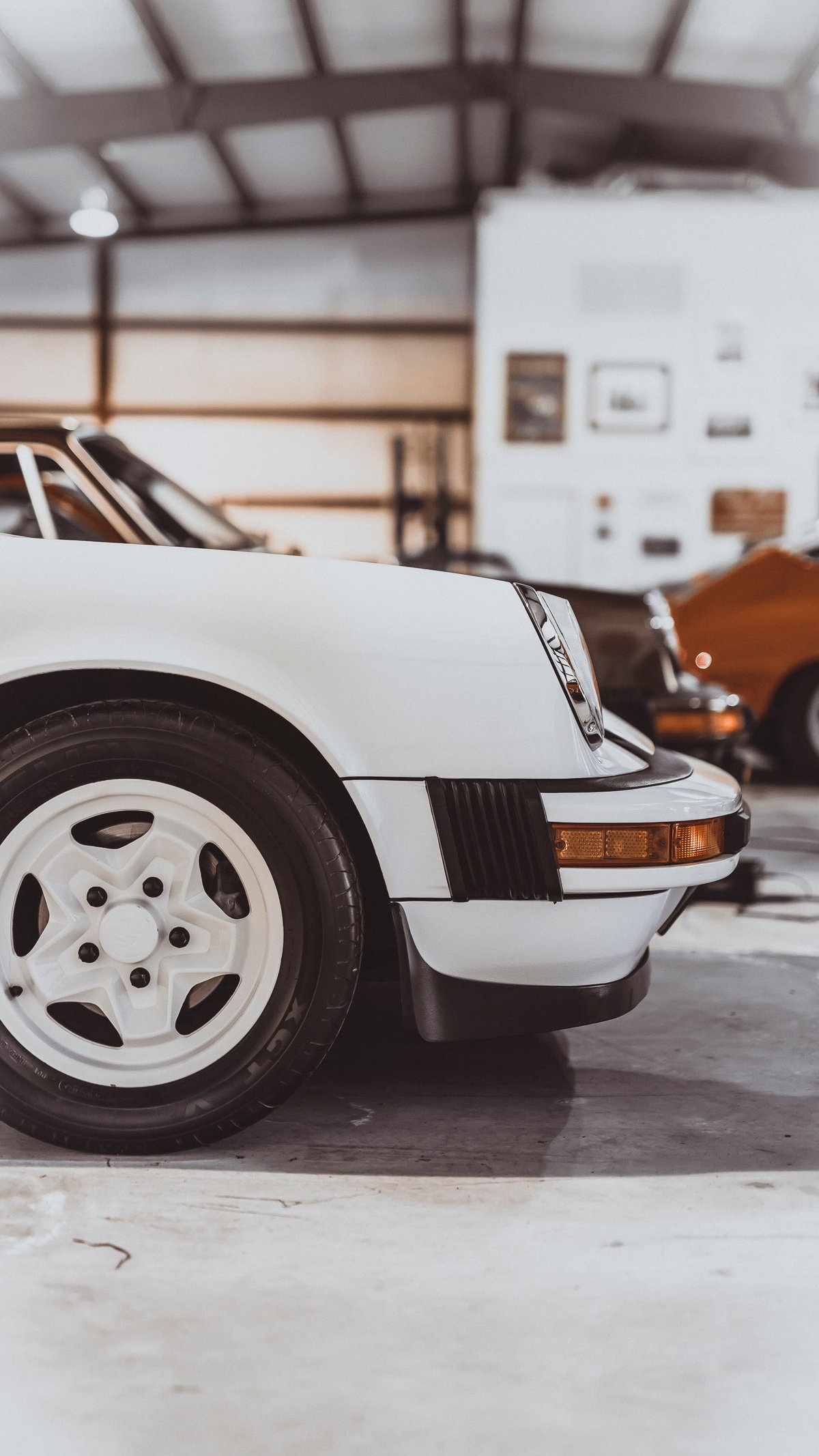 White Porsche 911 (G-series) front, featuring white 5-spoke wheels, black bumperettes, amber turn signal. Classic sports car.