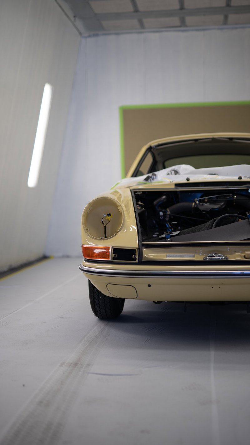 Beige Porsche 911 classic in a shop, left headlight bucket empty, frunk open. Displays vintage chrome bumper and amber turn signal.