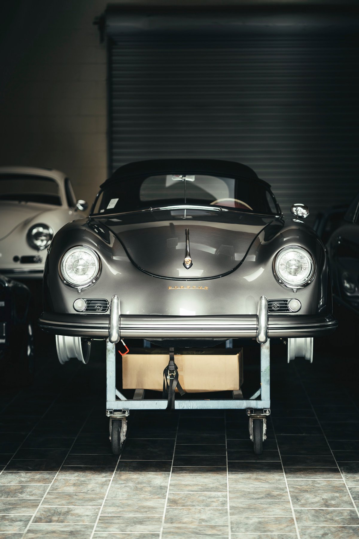 Front view of a metallic grey Porsche 356 Cabriolet on a workshop dolly, showcasing chrome bumpers, round headlights, and soft top.