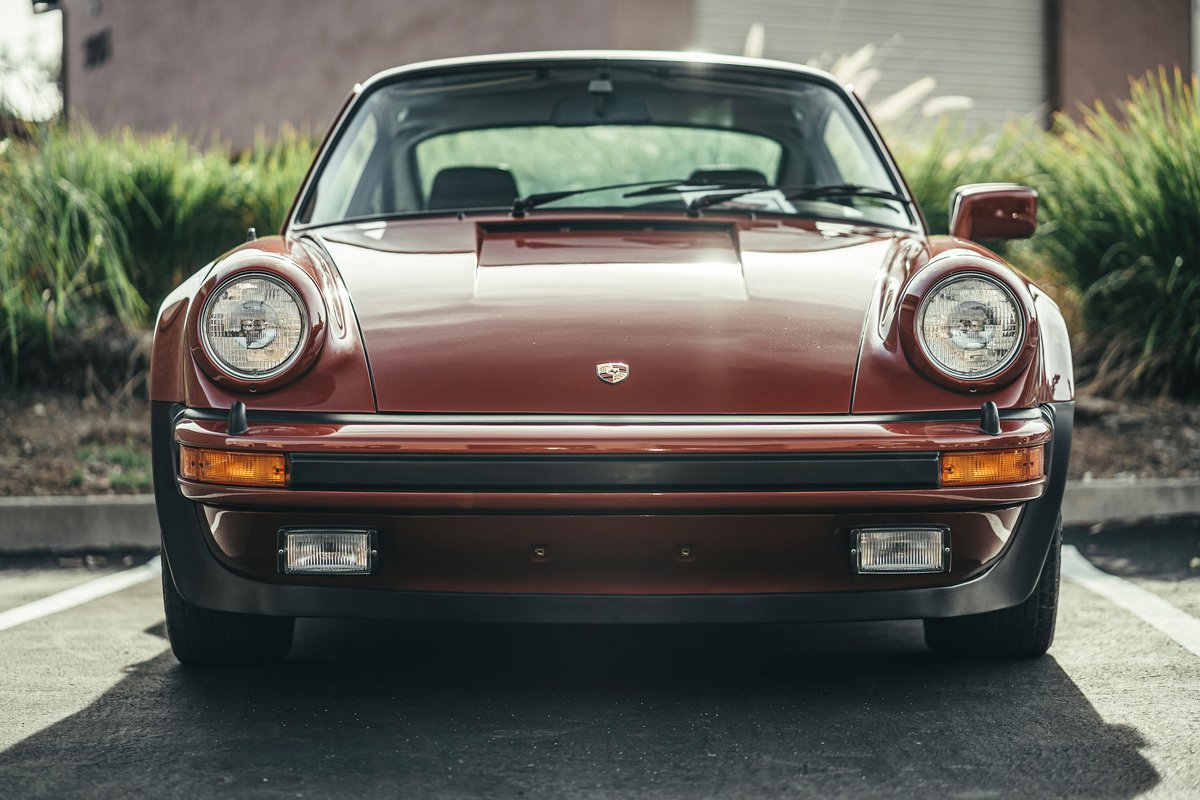 Front view of a deep red Porsche 911 G-series, showcasing its classic round headlights and US-spec rubber bumper trim.