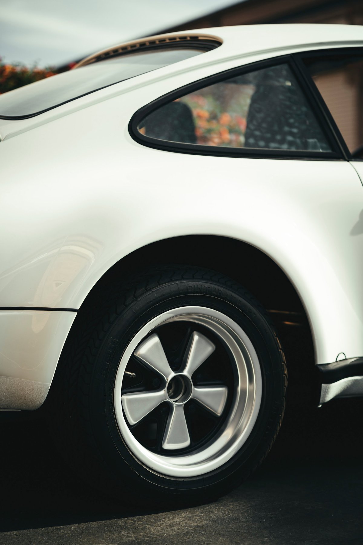 White classic Porsche 911 rear quarter panel, showcasing iconic polished Fuchs wheels, black window trim, and a sliding sunroof.