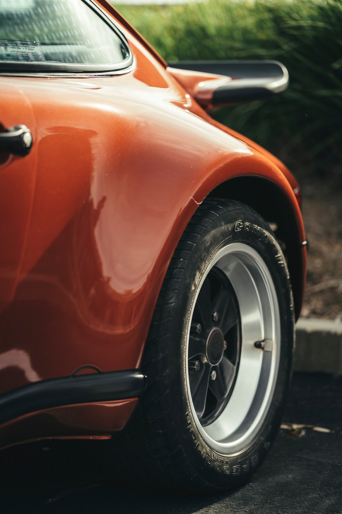 Metallic orange Porsche 911 G-series rear quarter. Features classic Fuchs wheels, BFGoodrich tires, and a ducktail spoiler.